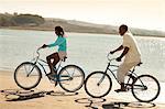 Smiling father and daughter riding their bikes alongside a beach.