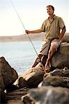 Smiling middle aged man sitting on a rock with a fishing rod.
