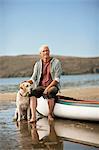 Happy senior man sitting on the edge of a canoe with his dog.