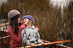 Young sisters smile at each other as they sit together in a wooden canoe among reeds on a lakeshore and pose for a portrait.