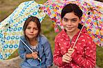 Two young girls sit on a fallen tree trunk while holding open umbrellas as they smile and pose for a portrait.