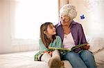 Grandmother reads a book with her young granddaughter as they sit together on a bed.