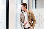 Young businessman holding mobile phone while looking through office window