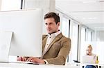 Confident young businessman working on computer with female colleague in background at office