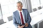 Thoughtful mature businessman holding digital tablet while leaning on conference table