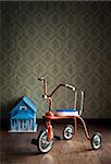 Vintage tricycle with doll house on background and vintage wallpaper.