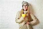 Happy Fashion Hipster Girl in Knitted Sweater and Beanie Hat with a Mug in Hands. Smiling Nice Woman at White Brick Wall Background. Winter or Autumn Warming Up Concept. Toned Photo with Copy Space.