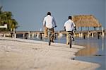 Mid-adult man and his son riding bicycles on a sandy beach near the water.