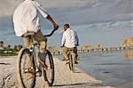 Smiling mid-adult man and his son riding bicycles on a sandy beach near the water.