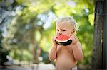 Portrait of a shirtless young toddler eating a large slice of watermelon outdoors.