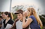 Teenage girl whispering in her boyfriend's ear while sitting outside with friends.