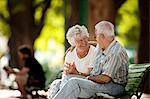 Senior woman smiles at a senior man with his arm around her shoulders as they sit together on a park bench.