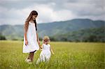 Girl and her younger sister wearing white sun dresses walk through a field.