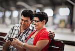 Mature couple sitting on an urban park bench, looking at something on a cell phone.