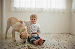 Young boy sitting on floor covered in dog biscuits with his dog.