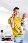 Mother holding baby daughter and talking on cell phone at laptop