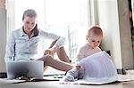 Curious baby daughter looking at paperwork next to mother working at laptop