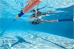 Amazing female swimmer swims underwater in the swimming pool outdoors. She wears a black-gray swimsuit with patterns, a white swim cap and swim glasses. Sunlight falls from above. Horizontal.