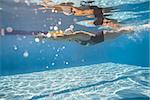 Active swimmer swims in crawl style underwater in the swimming pool outdoors. She wears swimsuit, swim cap and swim glasses. Her body reflected in water surface. Sunlight falls from above. Horizontal.