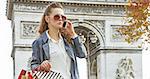 Stylish autumn in Paris. Full length portrait of modern woman in trench coat  with shopping bags in Paris, France using a smartphone