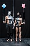 Nice couple of pole dancers holds their hands together next to a pylon on a dark background in the studio. They have body-art and wears black clothes. Girl and guy hold blue and pink balloons.