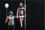 Sportive couple of pole dancers in the dark studio. Man holds a pylon with the left hand and holds a balloon in the right. Girl sits on the guy's left arm. They have a horrific body-art. Horizontal.