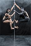 Photo of the couple of pole dancers with a horrific body-art in the dark studio with a cloud of a smoke. They are hanging on a pylon. Guy and the girl dressed in black sportswear. Vertical.