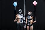 Gorgeous couple of pole dancers holds their hands together next to a pylon on a dark background in the studio. They have body-art and wears black clothes. Girl and guy hold blue and pink balloons.