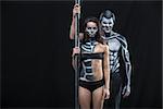 Grinning couple of pole dancers with horrific body-art stands next to a pylon on a dark background in the studio. They dressed in black sportswear and hold the right hands on the pylon. Horizontal.