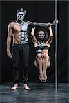 Perfect couple of pole dancers in the dark studio. Man holds a pylon with the left hand. Girl hangs with tighten knees on the guy's left arm. They wear black sportswear and have a horrific body-art.
