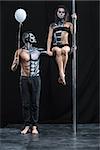 Strong couple of pole dancers in the dark studio. Man holds a pylon with the left hand and holds a balloon in the right. Girl sits on the guy's left arm. They have a horrific body-art. Vertical.