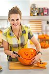 Frightful Treats on the way. Portrait of smiling modern housewife in the Halloween decorated kitchen carving a big orange pumpkin Jack-O-Lantern