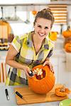 Frightful Treats on the way. Portrait of happy modern housewife in the Halloween decorated kitchen prepare big orange pumpkin for carving