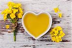 Rapeseed oil in heart shaped bowl and flowers on wooden background, top view.