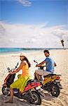 Smiling young couple having fun riding moped scooters along a tropical beach.