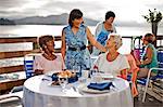 Two mature female friends out to lunch at a seaside restaurant share a laugh with an acquaintance who has come to say hello.