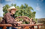 Teenage girl wearing a cowboy hat sits between the rusty railings of a corral and looks away pensively as she poses for a portrait.