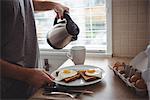 Man holding his breakfast plate while pouring hot water into mug in the kitchen