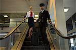 Pilot and air hostess with their trolley bags standing on escalator in the airport terminal