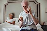 Worried senior man sitting in bedroom holding medicine and talking on mobile phone