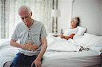 Worried senior man with hand on stomach sitting in bedroom