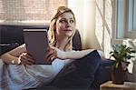 Thoughtful woman lying on sofa and holding digital table in living room at home