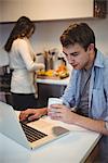 Man using laptop while woman working in background at kitchen