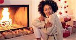 Woman smiles at camera while wearing sweater and seated in front of a raging fireplace and plastic white christmas tree