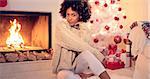 Lovely black woman in white leggings and sweater seating next to fireplace and christmas tree. She warming up by fire and woolen sweater.