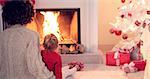 Mother and child warm up by the fireplace with a white christmas tree and presents nearby