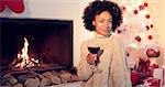 Pretty african american woman with huge afro haircut holds glass with red wine. She sitting next to fireplace and white christmas tree. She wearing winter woolen beige sweater.