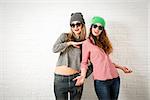 Two Happy Smiling Hipster Girls Posing at White Brick Wall Background. Trendy Casual Fashion Woman Outfit in Autumn or Spring. Teenage Female Friendship and Lifestyle. Toned Photo with Copy Space.