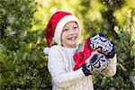 beautiful smiling boy in santa's hat, sweater and mittens holding nicely wrapped christmas gift by the tree or in the forest enjoying snowy cold winter weather, holiday or happiness concept