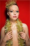 beauty portrait of pretty woman with creative make-up and golden hair-style accessory and tinsel on nude breast, she is posing on red background and looking in camera with charming eyes